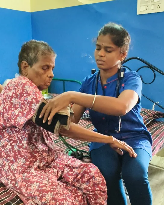 A nurse in blue adjusting a blood-pressure cuff on the arm of an older woman in a red patterned dress.