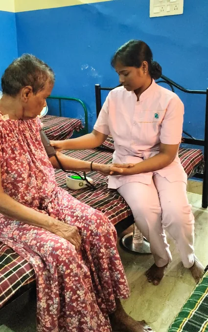 A nurse in pink seated beside an older woman and supporting a cuff on her arm.