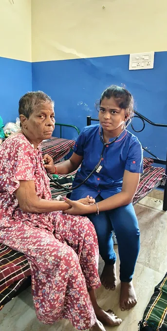 A nurse in blue seated beside an older woman in a patterned red dress.