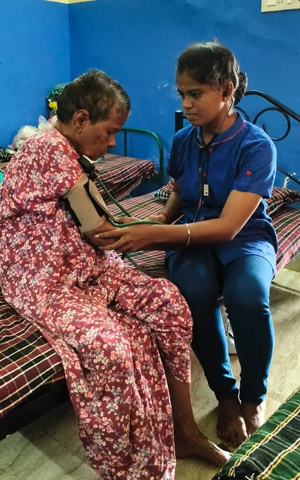 A nurse in blue using a stethoscope beside an older woman in a patterned red dress.