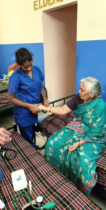 A nurse in blue assisting an older woman in a green sari with a blood-pressure cuff.