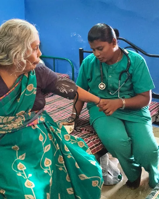 A nurse in green supporting an older woman in a green sari during a blood-pressure check.