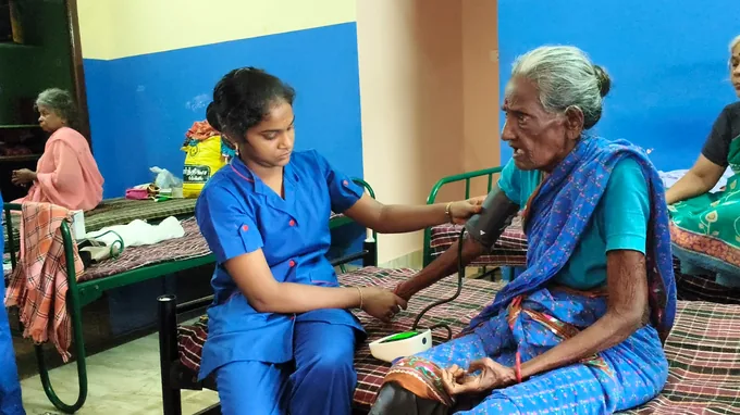 A nurse in blue supporting an older woman wearing a blue sari during a blood-pressure check.
