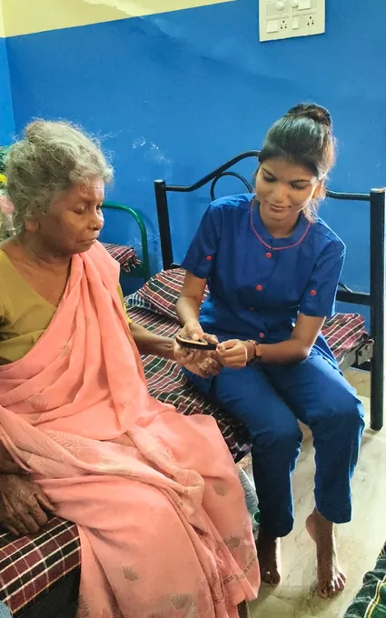 A nurse in blue carefully holding an older woman’s hand beside a small handheld device.