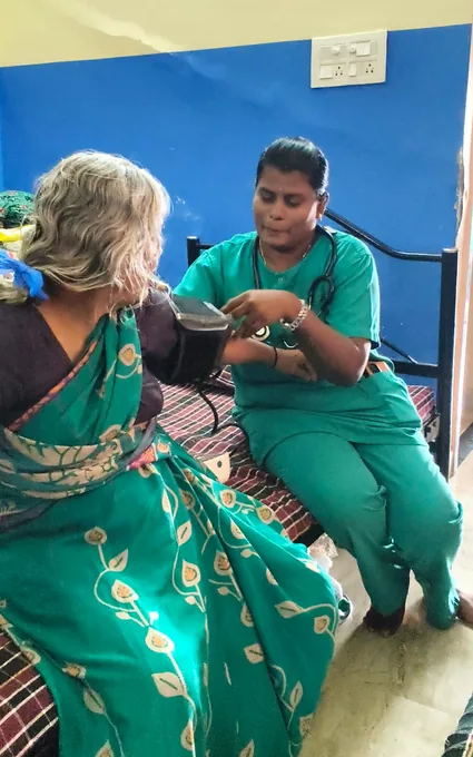 A nurse in green holding a blood-pressure cuff beside an older woman in a green sari.