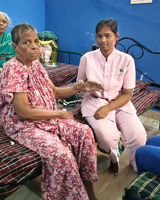 A nurse in pink seated beside an older woman in a red patterned dress and gently holding her hand.