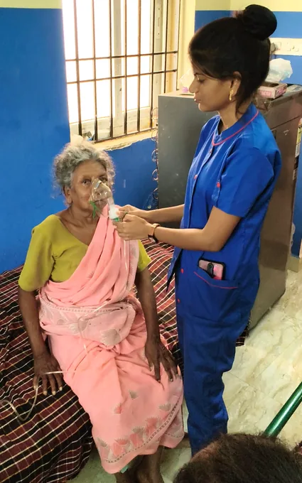 A nurse in blue standing beside an older woman in a pink sari and holding her hand.