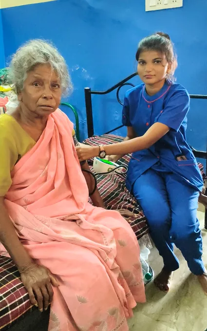 A nurse in blue beside an older woman in a pink sari, with a cuff around the woman’s upper arm.