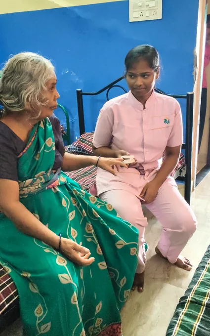 A nurse in pink using a stethoscope while seated beside an older woman in a green sari.