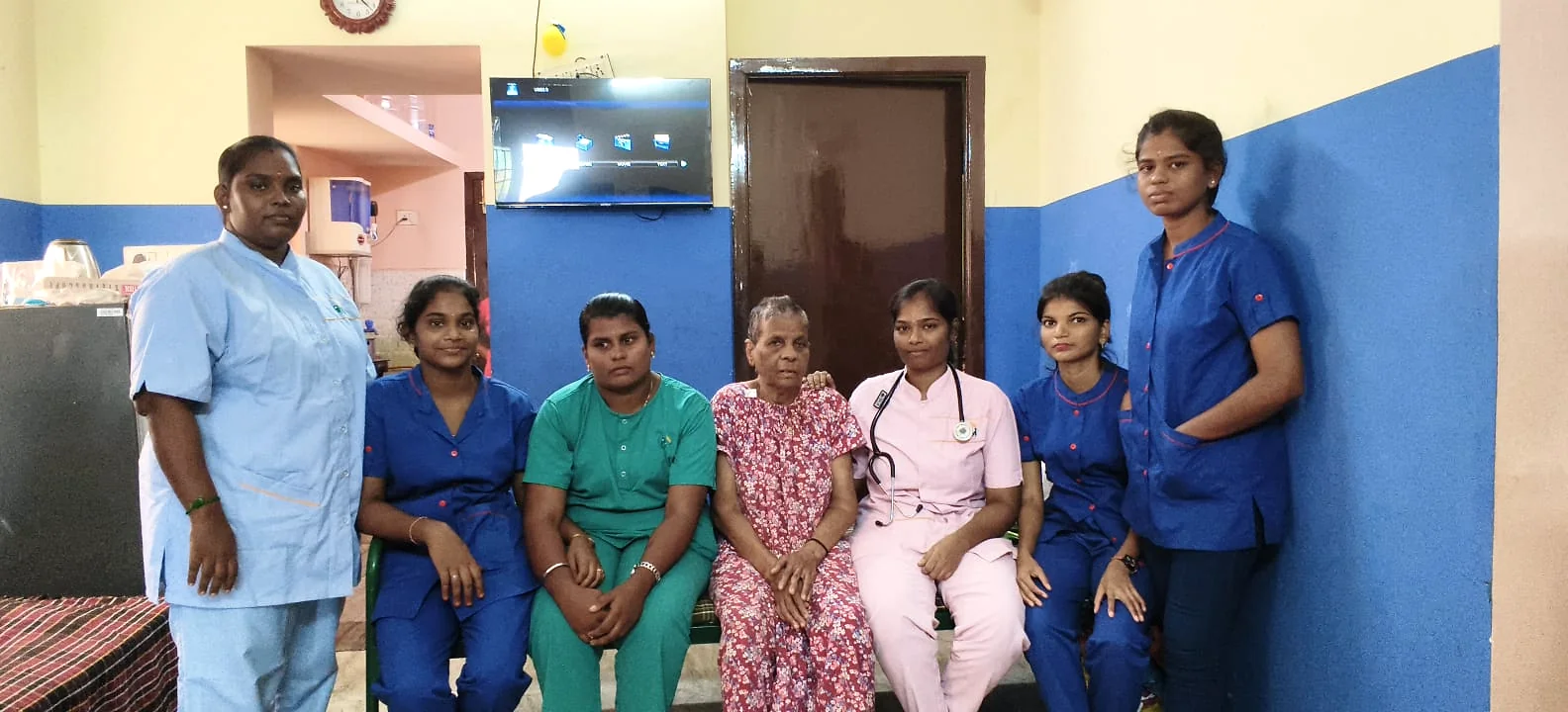 Six care-team members in blue, green and pink uniforms beside an older woman.
